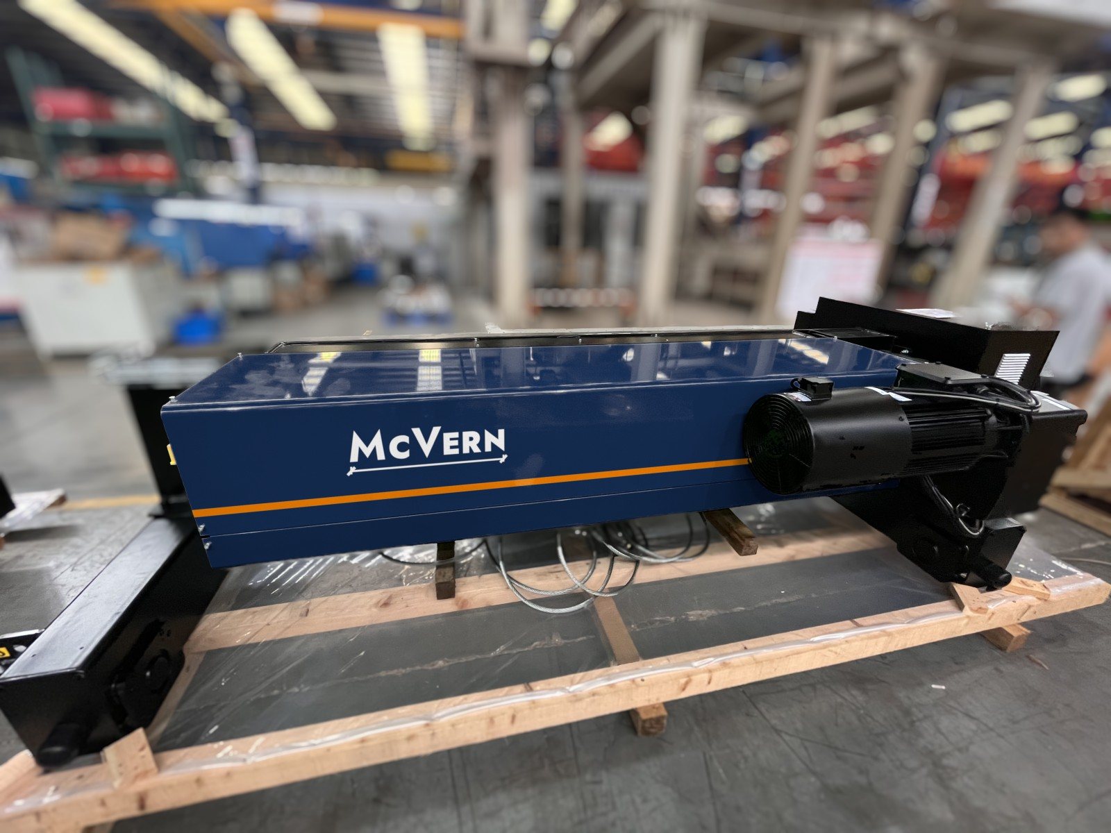 Wire-rope electric hoist unit in McVern deep navy with amber stripe and white logo, sharp against a softly blurred workshop